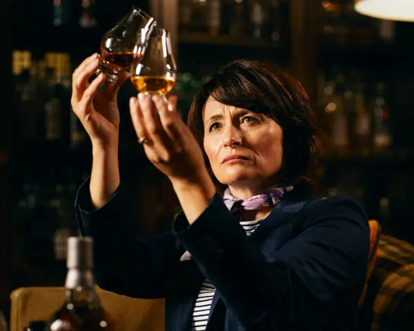 Rachel Barrie betrachtet konzentriert zwei Gläser Whisky, während sie in einer dunklen Bar sitzt.