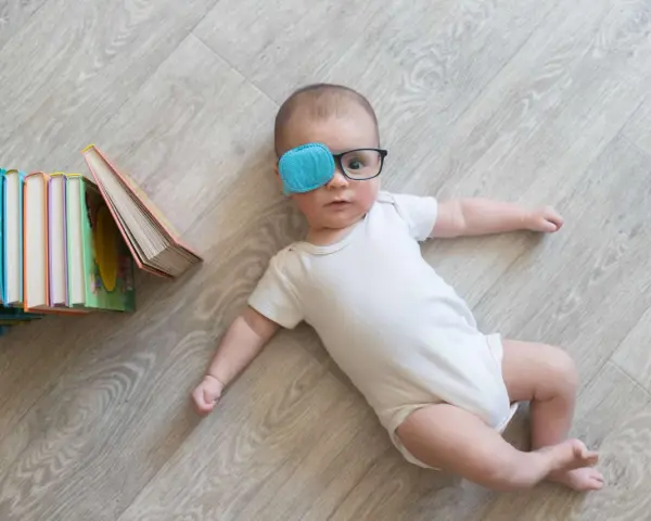 Bambino con occhiali e cerotto sull'occhio per correggere l'occhio ambliope, sdraiato vicino a una pila di libri colorati.