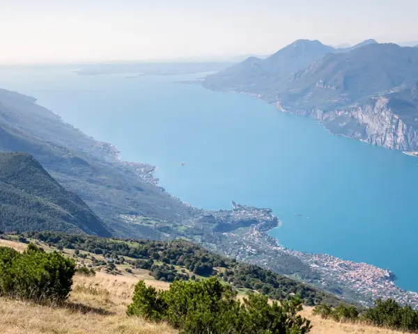 Panoramica mozzafiato del lago di Garda sponda veronese, con montagne verdi e un borgo pittoresco affacciato sull'acqua azzurra.