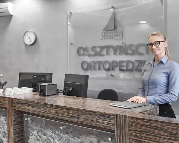 Uśmiechnięta recepcjonistka w okularach za ladą w gabinecie ortopedycznym.