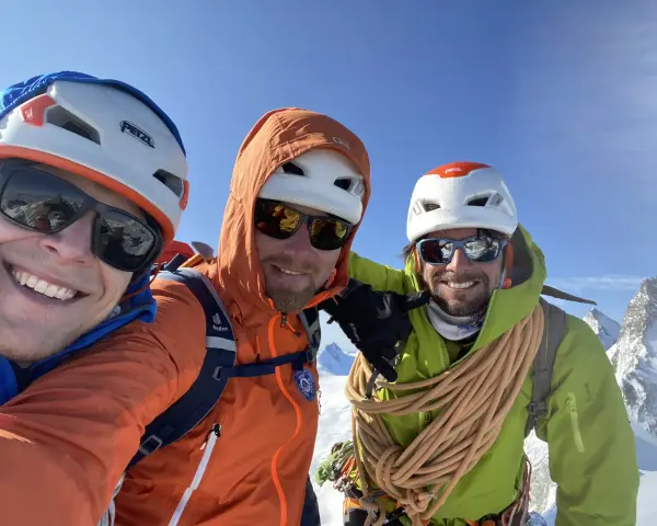 Trois alpinistes souriants au sommet, prêts à devenir guide de haute montagne à 40 ans. Corde et équipement de sécurité visibles.