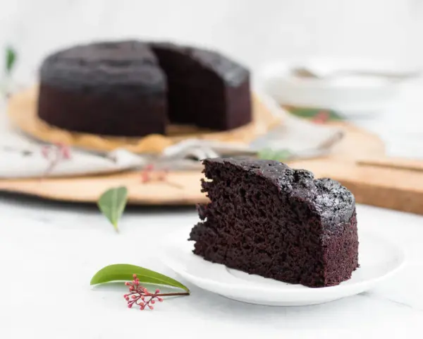 Ein saftiges Stück veganer Kuchen liegt auf einem Teller, daneben ein ganzer Schokoladenkuchen.