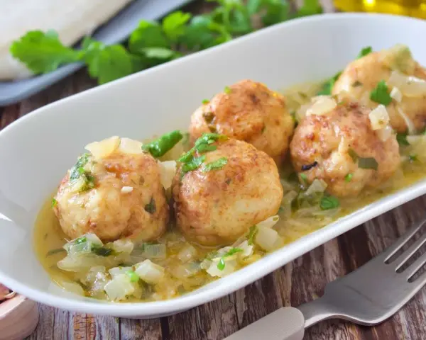 Cuatro albóndigas de merluza doradas en una salsa cremosa con cebolla y perejil, servidas en un plato blanco.