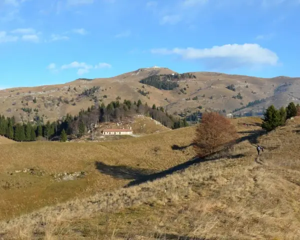 Sentieri facili sul Monte Grappa, con un escursionista che percorre un crinale erboso verso una casa isolata e la cima della montagna in lontananza.