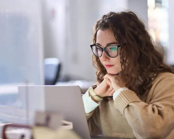 Donna con occhiali lavora al laptop, pensierosa. Potrebbe essere una dei nomadi digitali chi sono, che lavora da remoto.