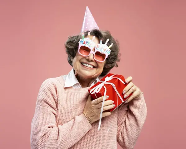 Uśmiechnięta starsza pani w okularach z napisem "Happy Birthday" i w czapce urodzinowej trzyma prezent. To dowcipne podziękowania za życzenia!