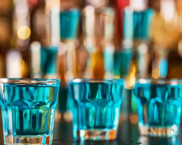 Trzy szklanki z niebieskim płynem, gotowe do wypicia jako kamikaze shot. W tle bar z butelkami.