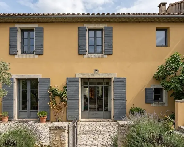 Façade d'une maison provençale à l'enduit à la chaux ocre, avec volets gris et végétation luxuriante.