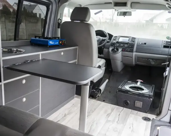 Wnętrze kampera w trakcie budowy: kuchenka turystyczna, rozkładany stolik, fotele i miejsce na toaletę.
