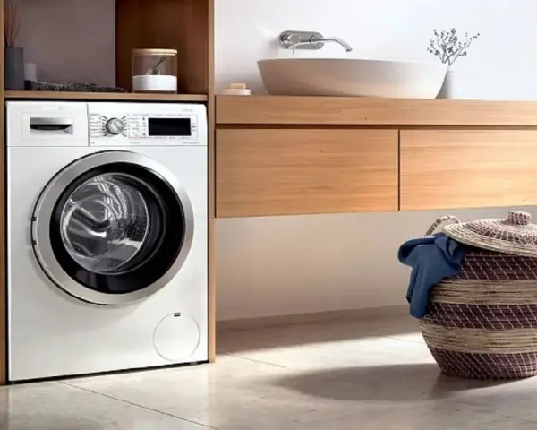 Lavadora blanca moderna lista para instalar en un baño con cesta de ropa sucia.