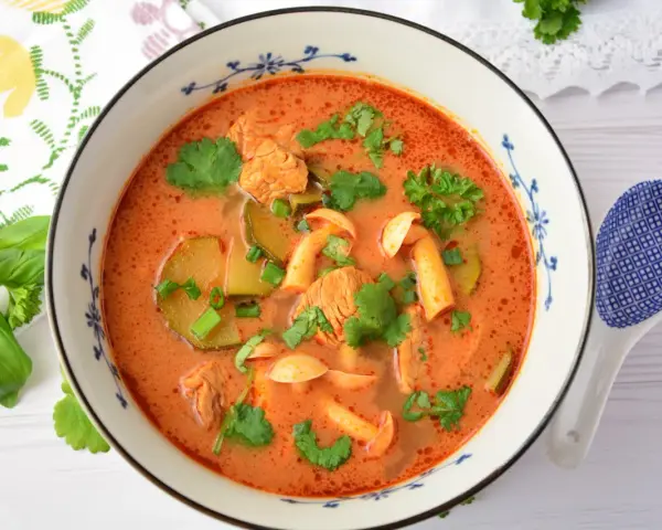 Aksamitny tom yum z kurczakiem, grzybami i kolendrą w misce z niebieskim wzorem. Obok leży łyżka.