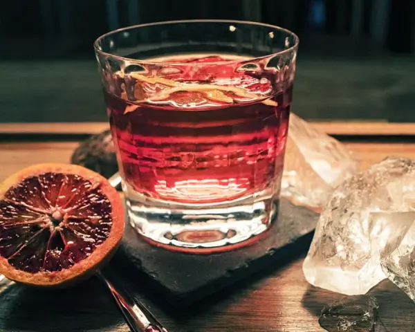 Ein Glas mit einem roten Drink, garniert mit Orangenschale, neben einer Blutorange und Eiswürfeln. Das ist Campari, ein italienischer Aperitif.