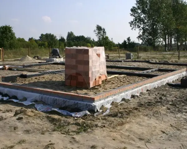 Stan zero budynku: fundamenty z cegły i betonu, piasek, folia izolacyjna, stos cegieł pod przykryciem.
