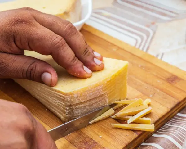 Mano cortando un bloque de queso amarillo en finas láminas. La corteza del queso se ve firme, indicando que se puede comer.