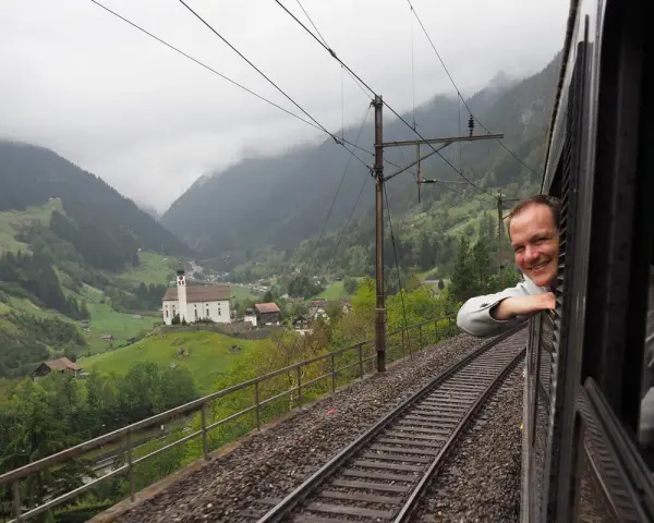 A smiling man leans out the window of the Gotthard Panorama Express, enjoying the stunning Swiss Alps scenery with a church nestled in the valley below.