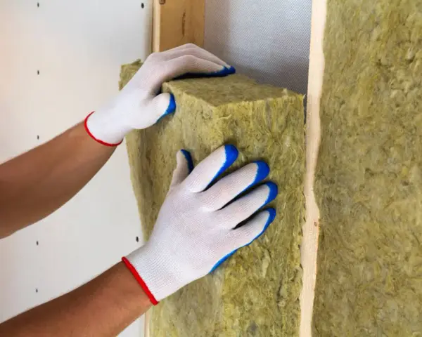 Mani guantate posizionano un pannello di lana di roccia, un materiale da **cemento isolante termico**, all'interno di una struttura in legno.