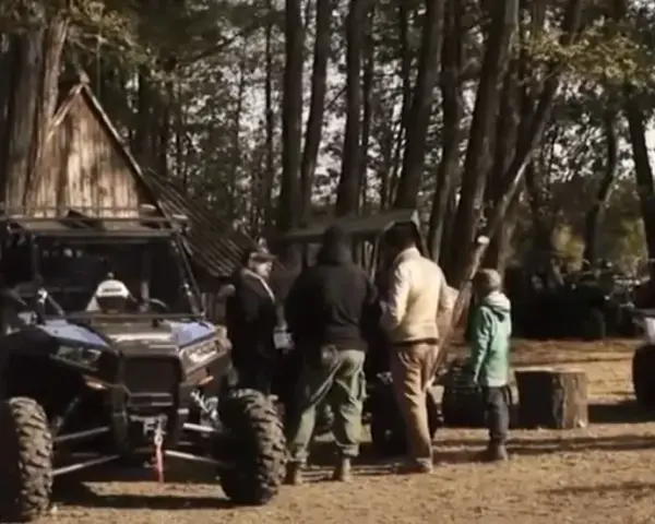 Grupa ludzi przy pojazdach terenowych podczas Polaris camping. W tle las i drewniana chata.