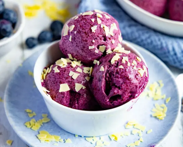 Drei Kugeln Blaubeer Eis, garniert mit weißen Schokostückchen, in einer weißen Schale auf einem blauen Teller.