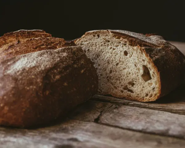 Ein rustikales, aufgeschnittenes sauerteigbrot ohne kneten liegt auf einer Holzoberfläche. Die Kruste ist dunkelbraun und knusprig, das Innere luftig und porös.