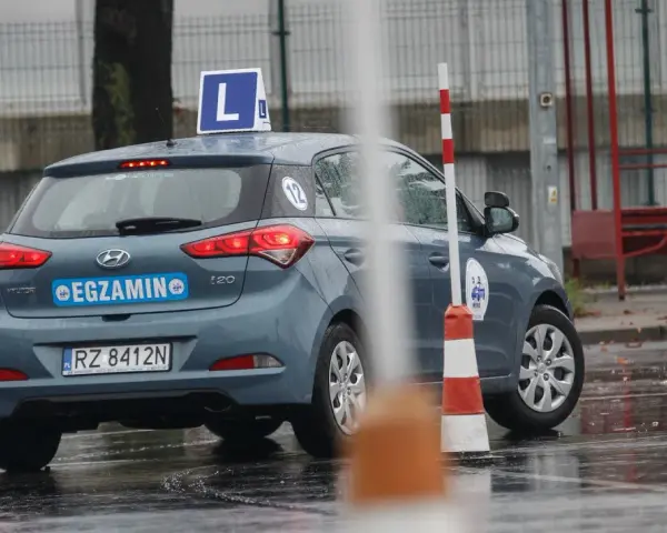 Samochód egzaminacyjny z literą "L" na dachu i napisem "EGZAMIN" na tylnej klapie, otoczony pachołkami na mokrym placu.