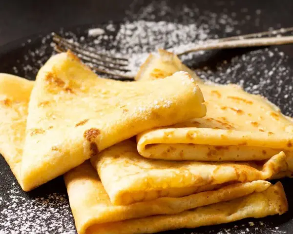 Deliciosos crepe en francés, doblados y espolvoreados con azúcar glas sobre un plato negro.