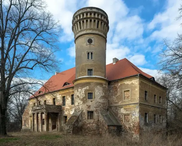 Zniszczony pałac w Osetnie z charakterystyczną wieżą i czerwoną dachówką, otoczony drzewami.
