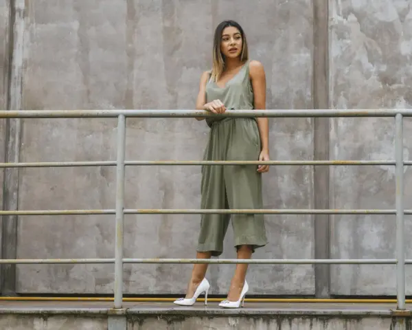 Kobieta w zielonym kombinezonie i białych szpilkach stoi przy metalowej balustradzie na tle betonowej ściany.