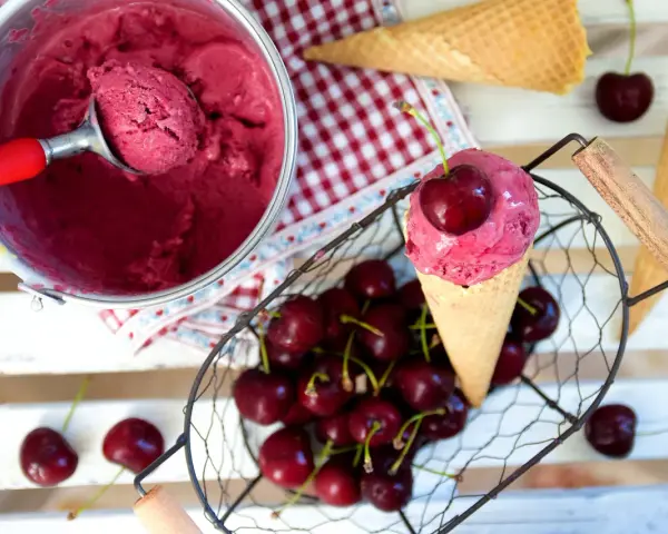 Ein Eisbecher mit Kirscheis, eine Kugel in einer Waffel, frische Kirschen und leere Waffeln.