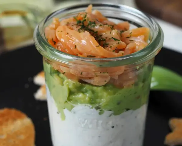 Leckerer Lachs Avocado Vorspeise im Glas, geschichtet mit cremiger Sauce und garniert mit Dill. Dazu geröstete Brotscheiben.