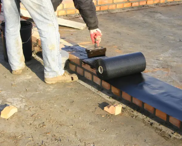 Pracownik kładzie papę na murze, szukając alternatywy dla dysperbitu. Czarna rolka materiału hydroizolacyjnego jest rozwijana na mokrej powierzchni.