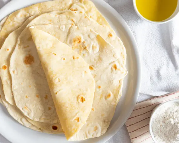 Piatto di piadine senza strutto, morbide e dorate, pronte per essere farcite. Accanto, una ciotolina d'olio e una di farina.