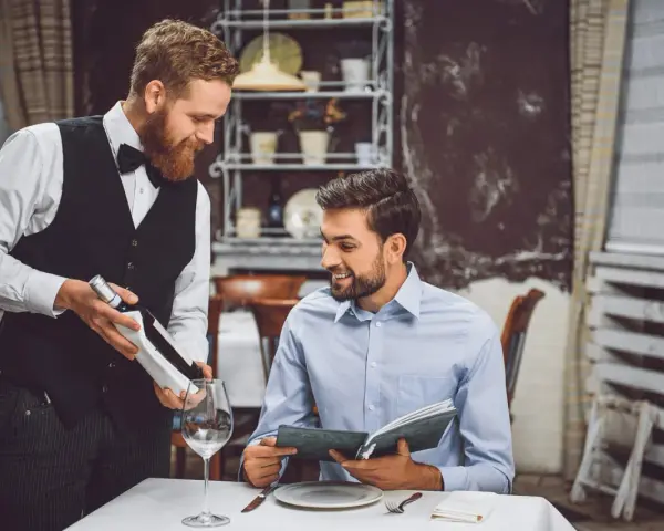 Camarero atento presenta una botella de vino a un cliente que revisa el menú. Demuestra las cualidades de un camarero: servicio, amabilidad y conocimiento.