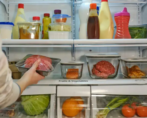 Mano colocando carne picada envasada en un refrigerador lleno de alimentos.
