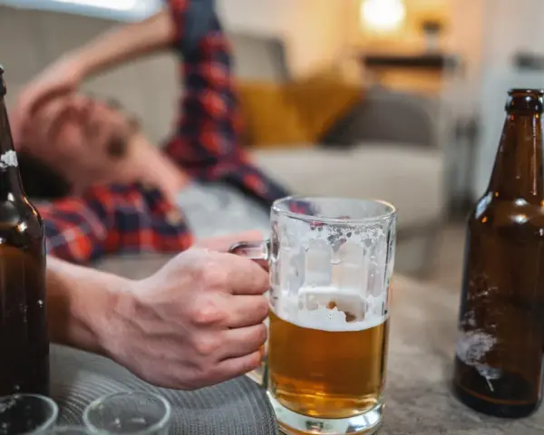 Mężczyzna leży na kanapie, trzymając kufel piwa. Wokół puste butelki, sugerujące nagły spadek tolerancji na alkohol.