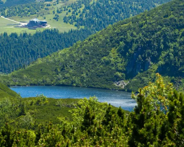Widok na górskie jezioro i schronisko Samotnia w Karkonoszach. Otoczone zielenią, lasem i skalistymi zboczami.