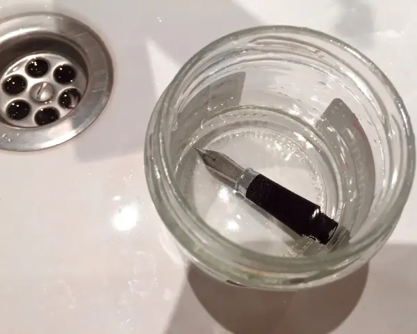 Pluma estilográfica sumergida en un frasco de vidrio con agua, junto a un desagüe. Un paso para aprender como limpiar una pluma.