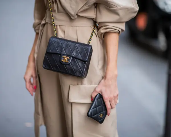 Elegancka kobieta w beżowym płaszczu prezentuje klasyczną, pikowaną torebkę Chanel vintage z złotym łańcuszkiem i logo.