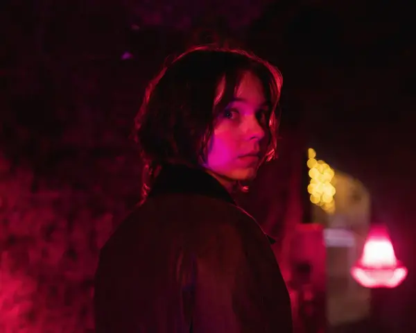 Młoda kobieta z zaciekawieniem spogląda przez ramię w mrocznym, neonowym otoczeniu. Czy to scena z nowego serialu Netflix?