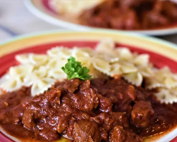 Gulasz z indyka z warzywami, podany z makaronem w kształcie motylków, ozdobiony natką pietruszki. Pyszne danie na talerzu.