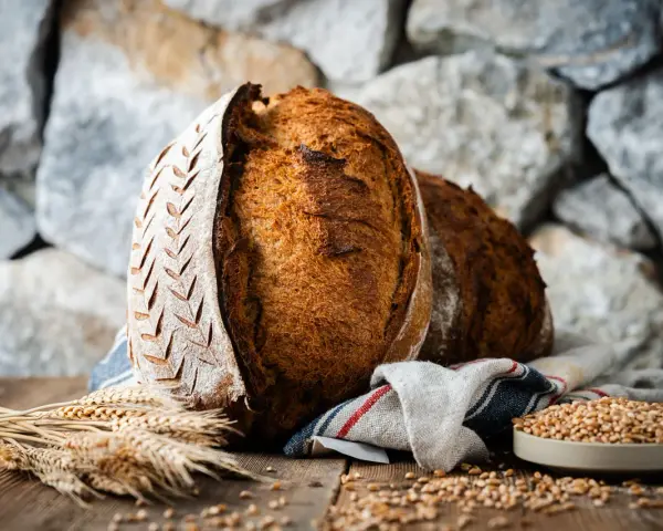 Zwei rustikale Brote, eines mit eingekerbtem Muster, liegen auf einem Holztisch. Weizenähren und eine Schale voller Körner deuten auf ein köstliches Ruchbrot Rezept hin.
