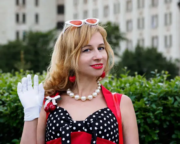Una mujer con guantes blancos saluda, luciendo un vestido de lunares y cerezas. ¡Buenos días, señorita en francés!