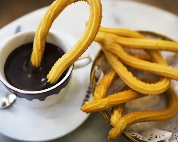 Churros sin gluten dorados y crujientes, uno sumergido en chocolate caliente, otro en una cesta.