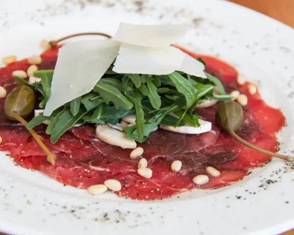 Delicato carpaccio di manzo con rucola, scaglie di parmigiano, pinoli, capperi e funghi.