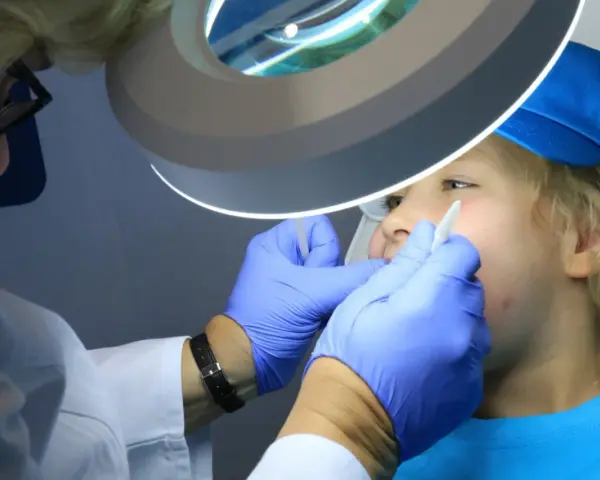 Dentysta w rękawiczkach bada usta dziecka pod lampą powiększającą.