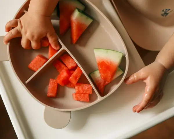 Kleine Hände greifen nach saftigen Wassermelone-Stücken auf einem Teller. Ein süßer Wassermelone-Baby-Moment.