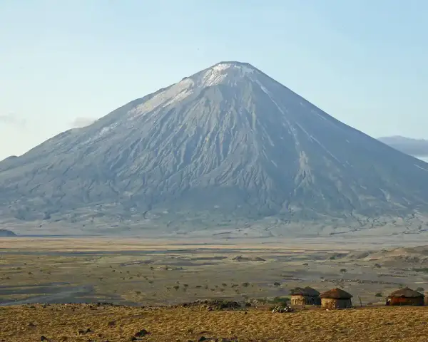 Ol Doinyo Lengai, wulkan w Tanzanii, góruje nad sawanną z kilkoma tradycyjnymi chatami Masajów.