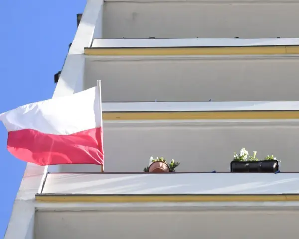 Prawidłowa flaga Polski powiewa na tle błękitnego nieba, obok balkonów z kwiatami.