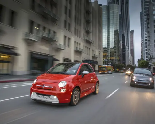 Czerwony Fiat 500e jedzie ulicą w mieście, w tle wieżowce i autobus.