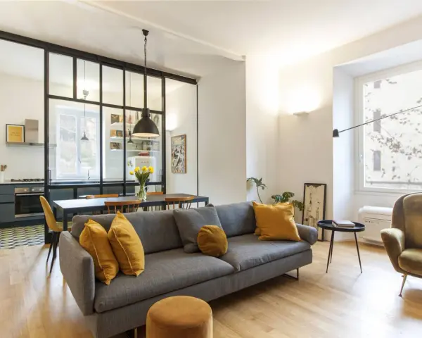 Ampia zona giorno con divano grigio e poltrona verde, separata dalla cucina da una moderna mezza parete divisoria cucina soggiorno in ferro e vetro.