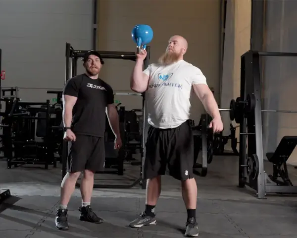 Man med skägg utför en kettlebell press med en blå kettlebell. En annan man står bredvid och tittar.
